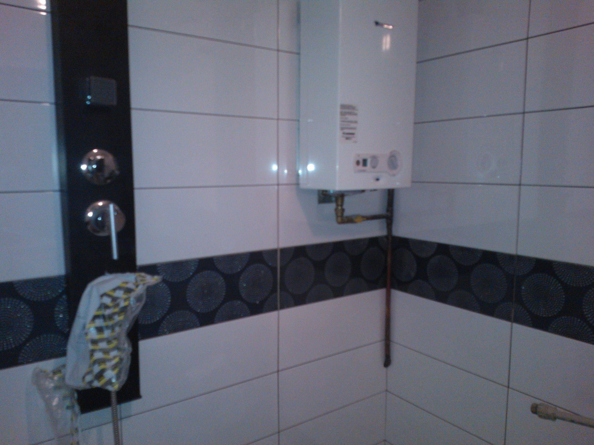 Biały piec gazowy zamontowany na ścianie w łazience z biało-czarnymi płytkami, widoczny panel prysznicowy z chromowanymi elementami, całość w trakcie montażu, prysznic zabezpieczony folią.