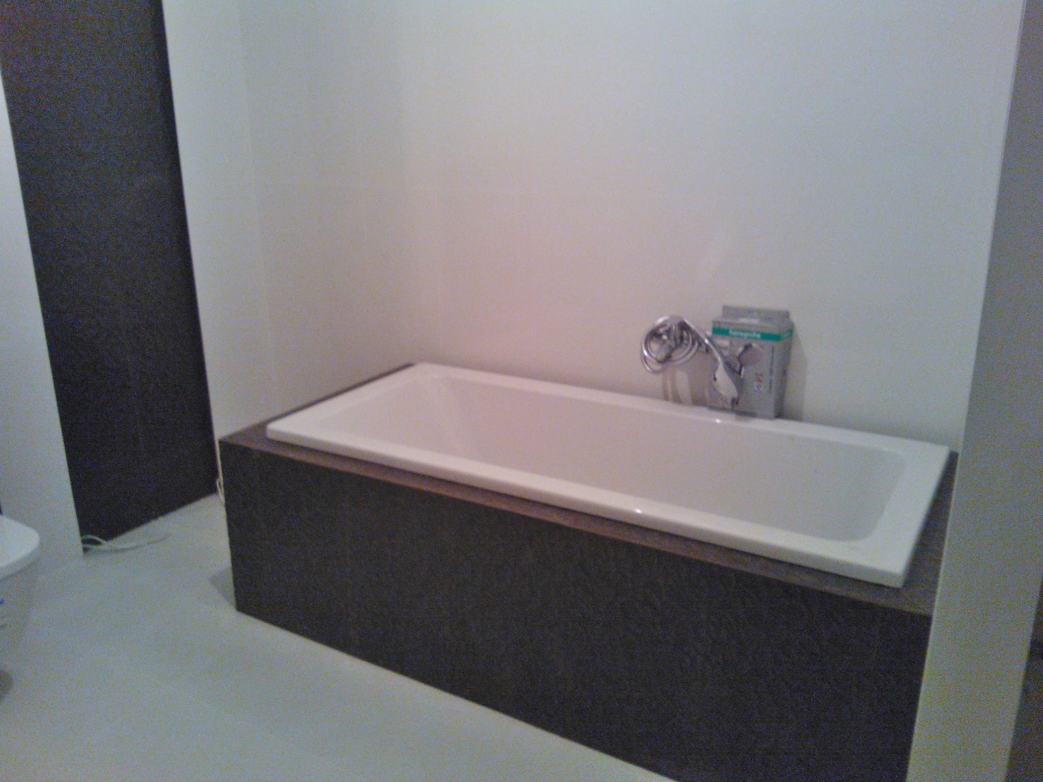 Biała wanna obudowana ciemnobrązową mozaiką, bateria umieszczona na wannie, w tle biała ściana i fragment toalety.