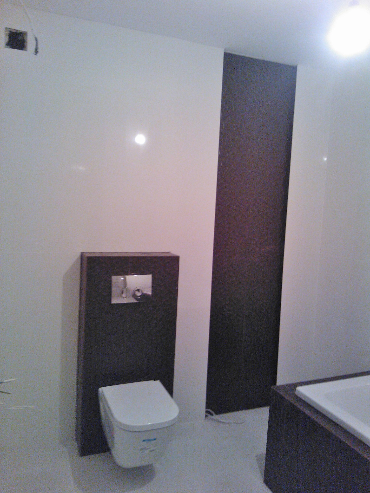 Nowoczesna łazienka z podwieszaną toaletą, zabudowaną spłuczką i akcentem w postaci ciemnej, pionowej płytki na ścianie, uzupełniona narożną wanną.