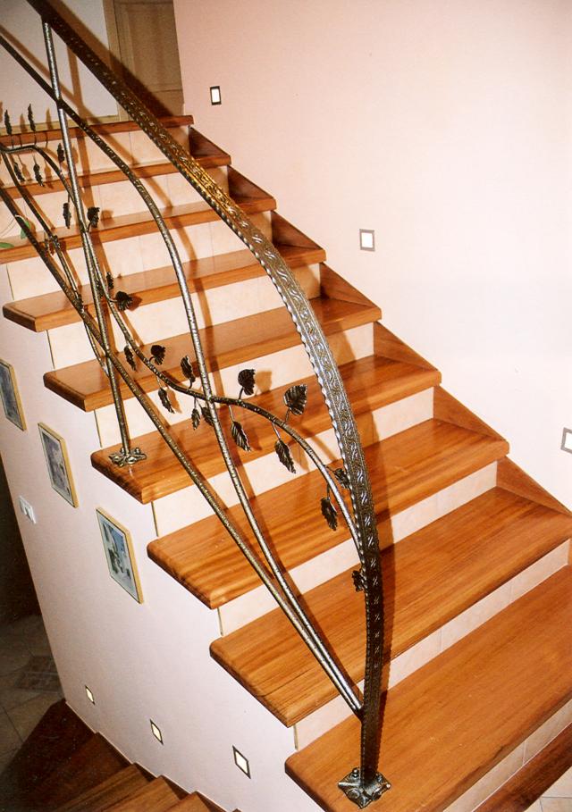 Drewniane schody z metalową, zdobioną balustradą z motywem liści, oświetlone punktowymi lampami w ścianie i pod stopniami.