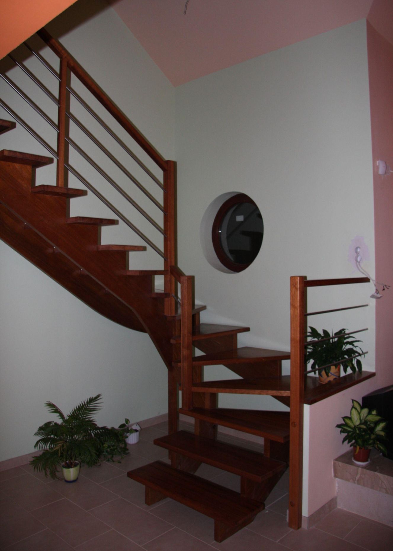 Drewniane schody dwubiegowe z balustradą z poziomych, metalowych prętów, okrągłe okno w ścianie, rośliny doniczkowe na półce i pod schodami.