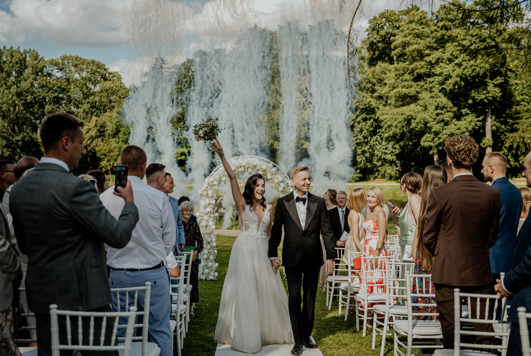 Uśmiechnięta para młoda, trzymając się za ręce, przechodzi alejką między gośćmi, za nimi dekoracyjny łuk z kwiatów i fontanna dymna.
