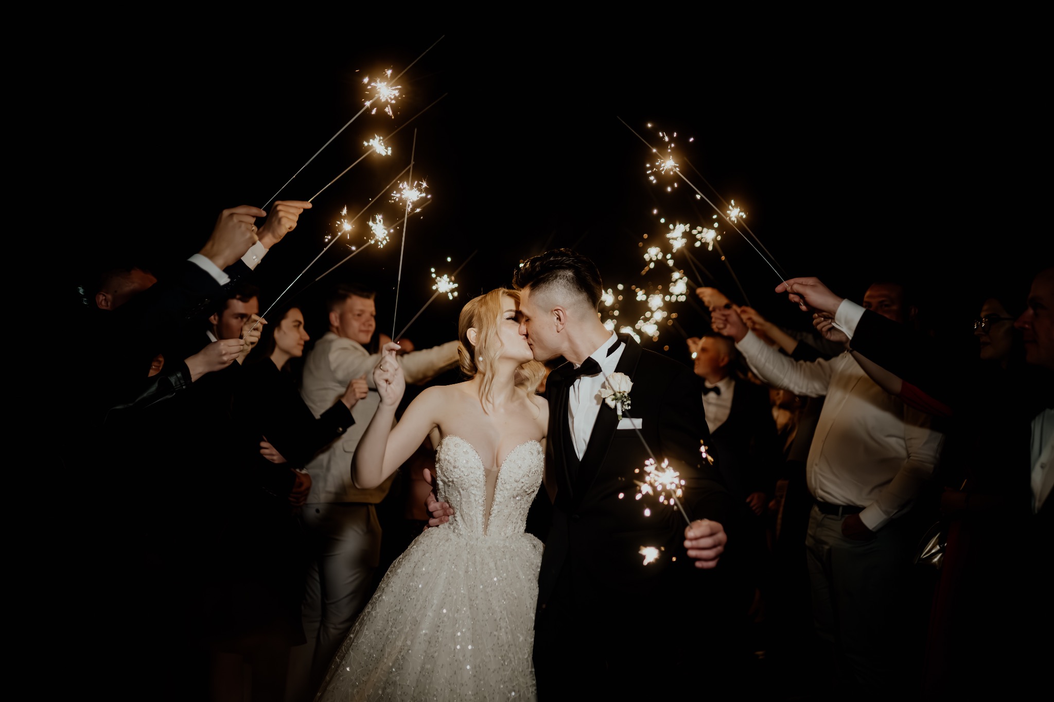 Pocałunek pary młodej otoczonej gośćmi z zimnymi ogniami na tle nocnego nieba, panna młoda w sukni z głębokim dekoltem, pan młody w smokingu z białą butonierką.