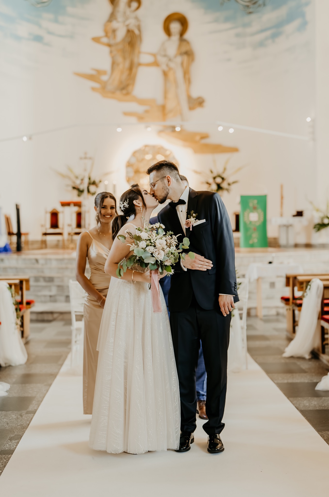 Pocałunek nowożeńców w kościele, panna młoda trzyma bukiet z różową wstążką, druhna w złotej sukni w tle, obraz religijny na ścianie.