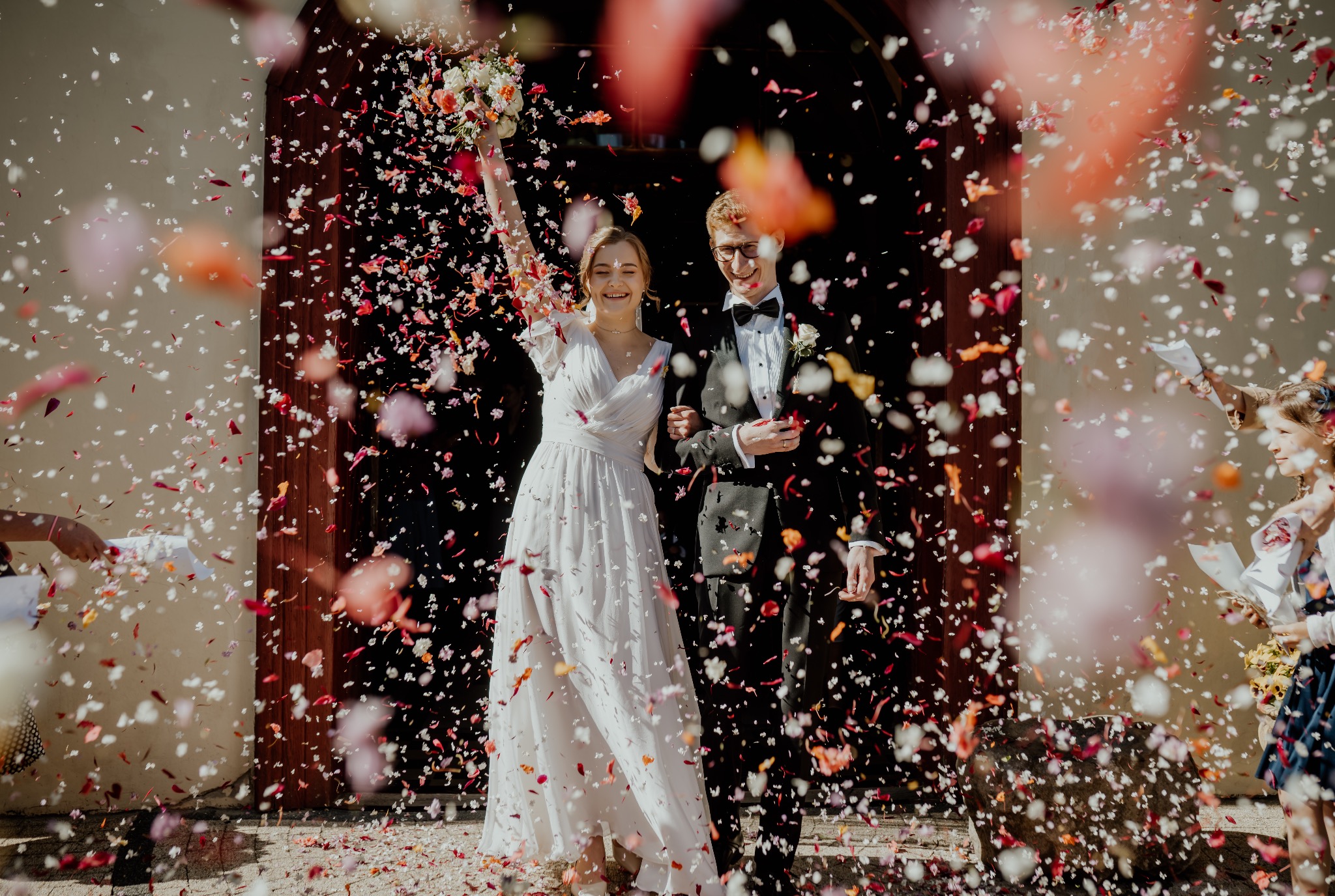 Uśmiechnięta para młoda w otoczeniu konfetti przed ciemnymi drzwiami, panna młoda trzyma bukiet kwiatów w uniesionej ręce.