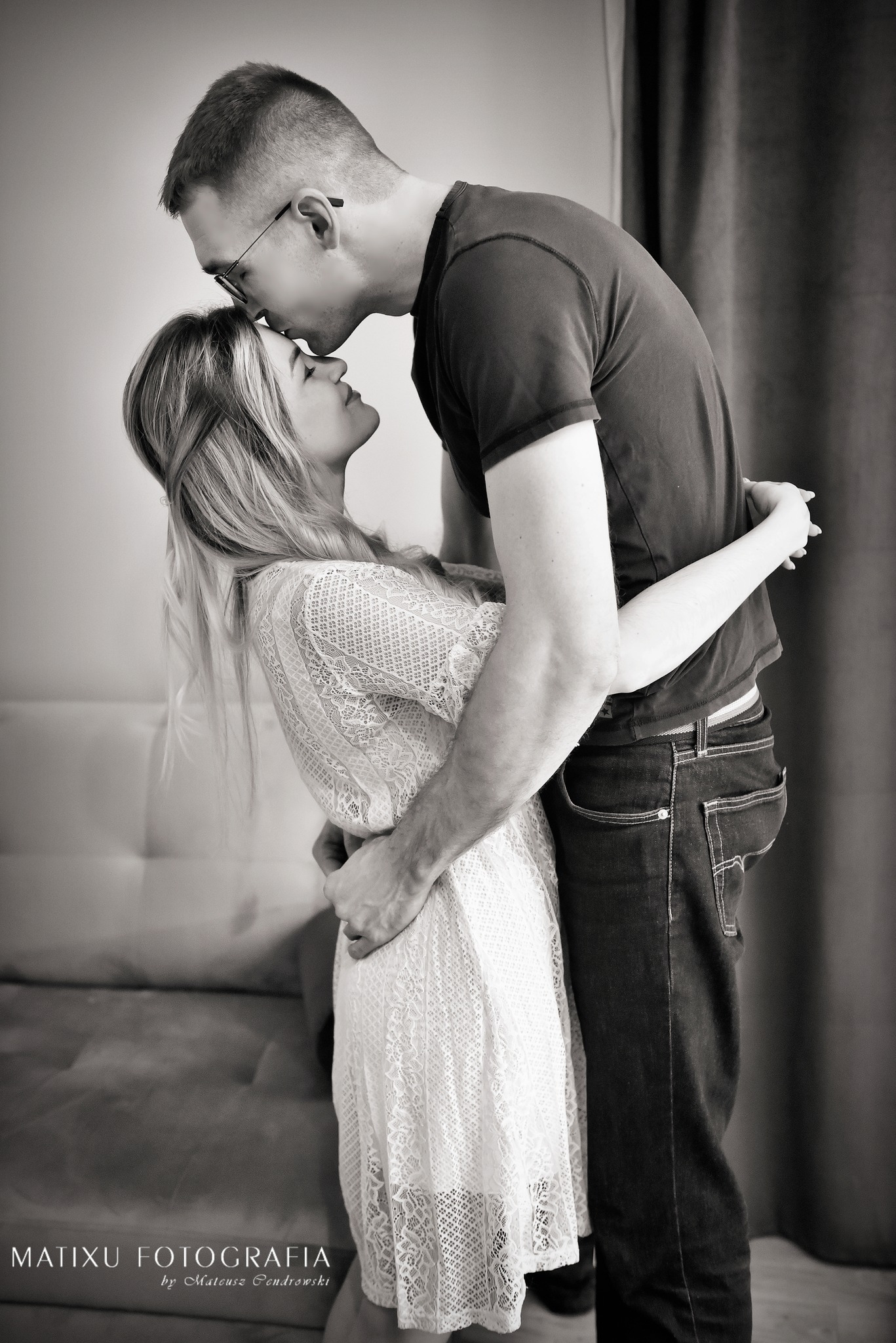 Czarno-białe zdjęcie obejmującej się pary; mężczyzna w okularach całuje kobietę w koronkową sukienkę w czoło.