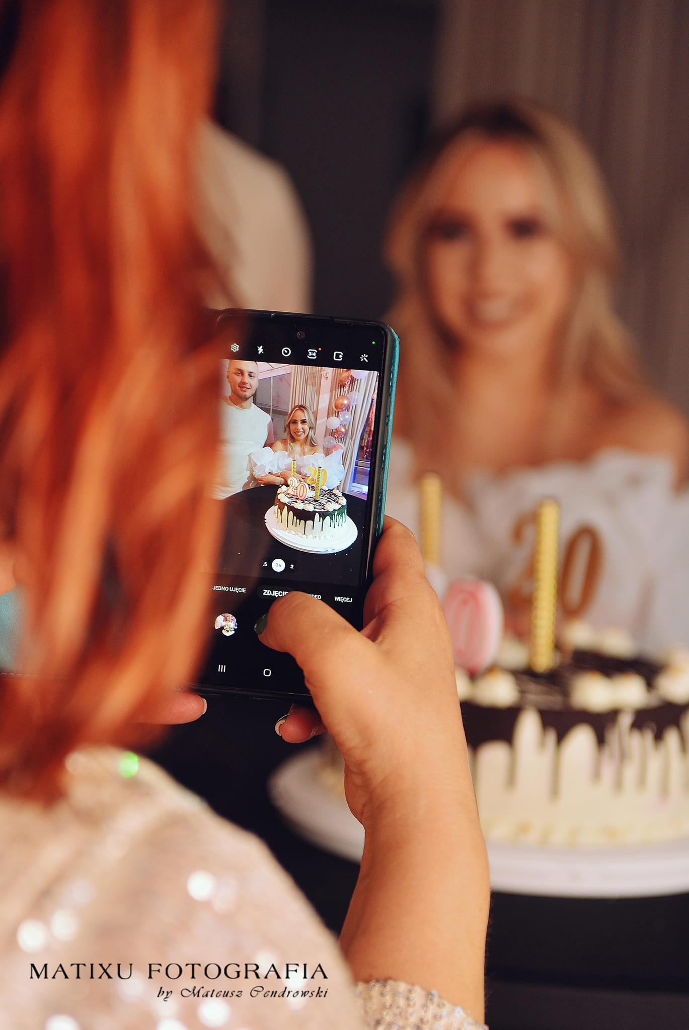 Osoba fotografuje telefonem parę z tortem urodzinowym '20' w Sztumie, widoczne świeczki i dekoracje balonowe w tle.