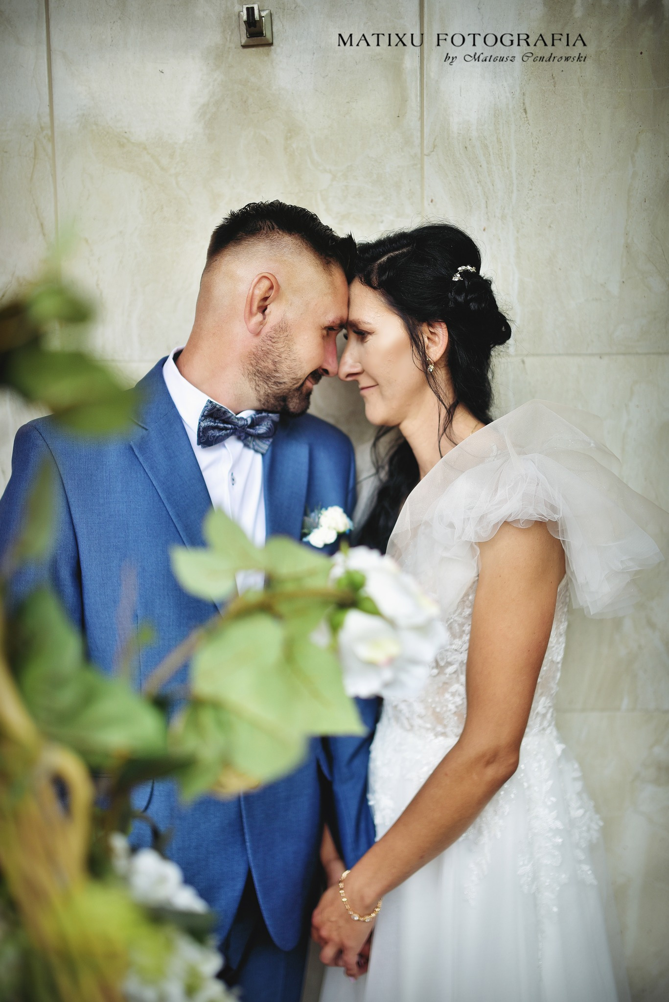 Para młoda dotykająca się czołami w zbliżeniu, on w błękitnym garniturze i muszce, ona w białej sukni z bufiastymi rękawami, na tle marmurowej ściany, częściowo zasłonięci przez rośliny.