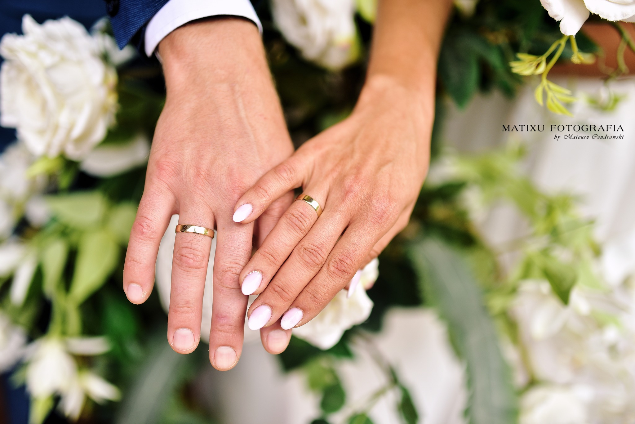 Dłonie pary młodej z złotymi obrączkami na tle białych kwiatów, zbliżenie na dłoń kobiety z delikatnym zdobieniem na paznokciu.