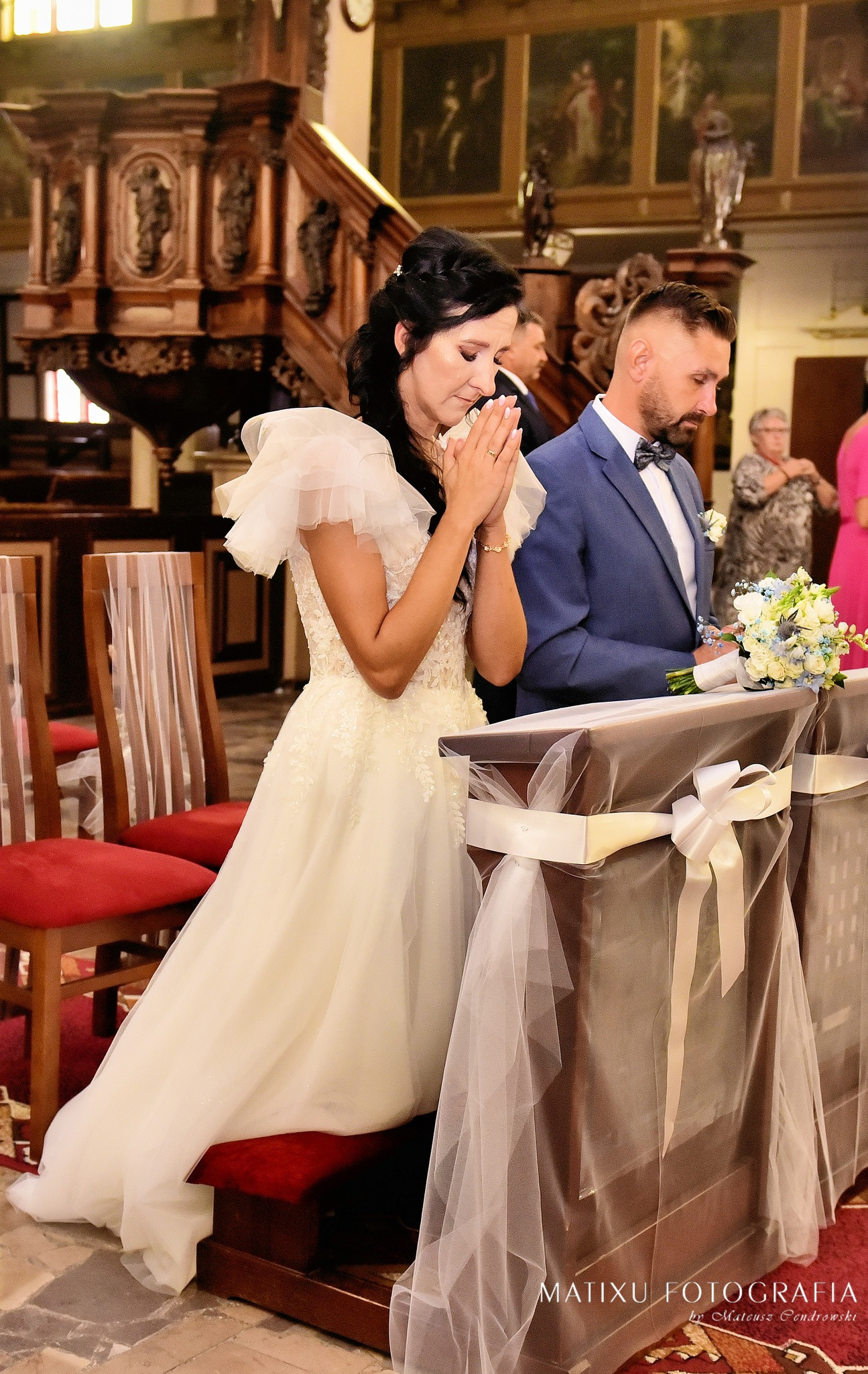 Panna młoda w białej sukni z bufkami klęczy przed ołtarzem w kościele, modląc się ze złożonymi dłońmi, obok stoi pan młody w niebieskim garniturze z bukietem.