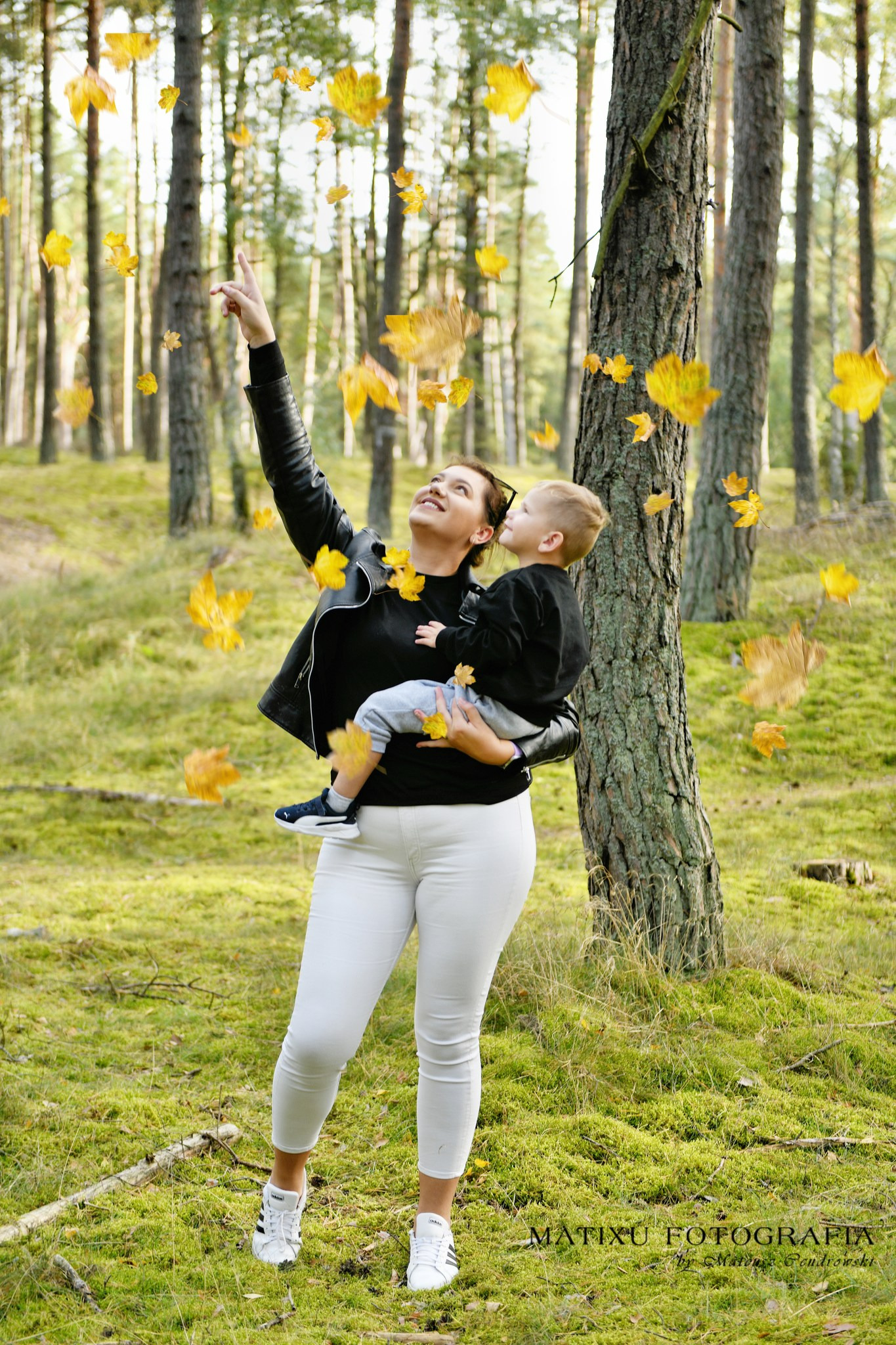 Kobieta w czarnej kurtce trzyma dziecko na rękach w lesie, liście opadają, kobieta wskazuje palcem w górę, leśne tło.