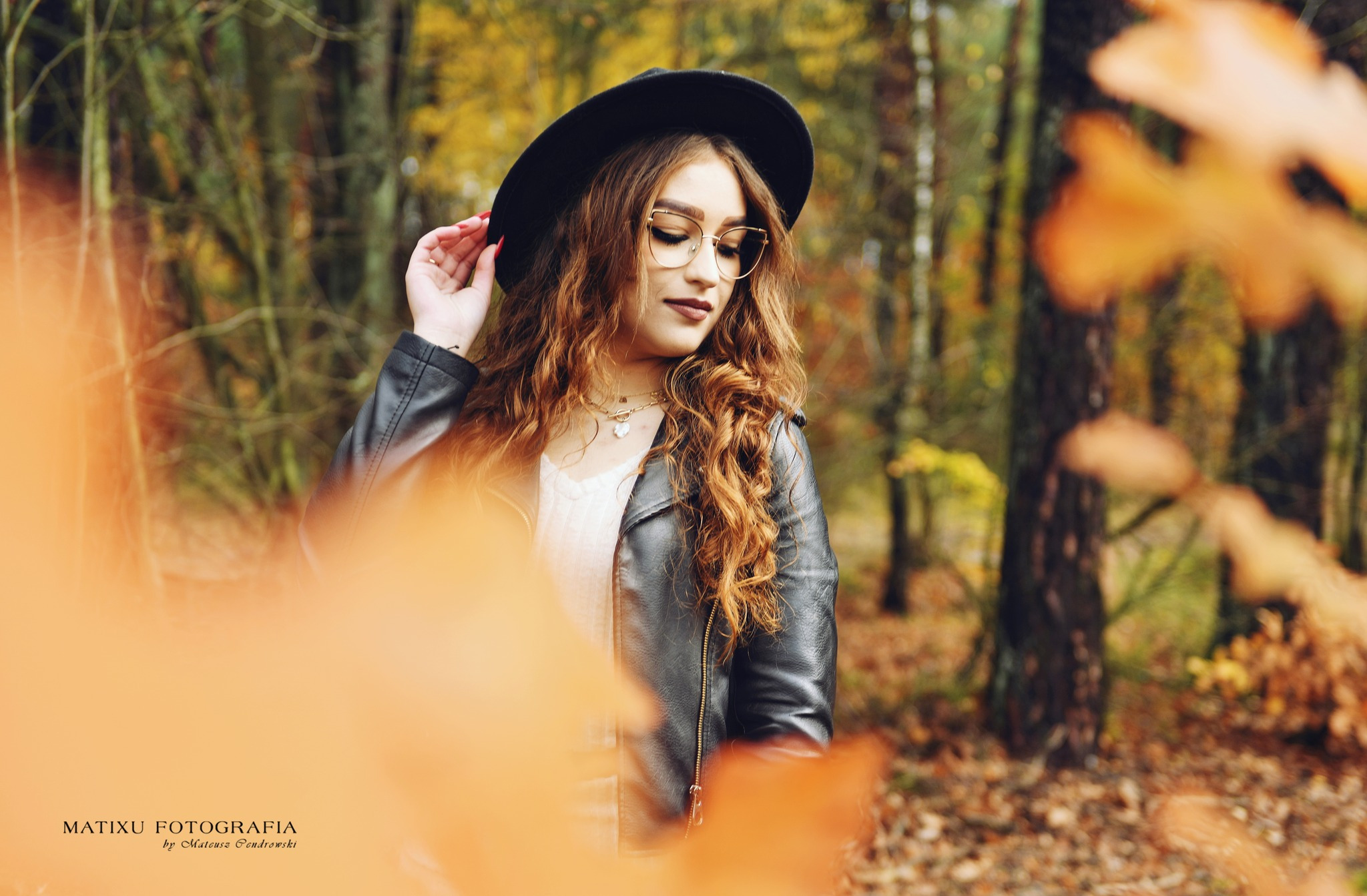 Portret młodej kobiety w kapeluszu i okularach, z rudymi kręconymi włosami, w skórzanej kurtce, pozującej na tle jesiennego lasu, z liśćmi na pierwszym planie.
