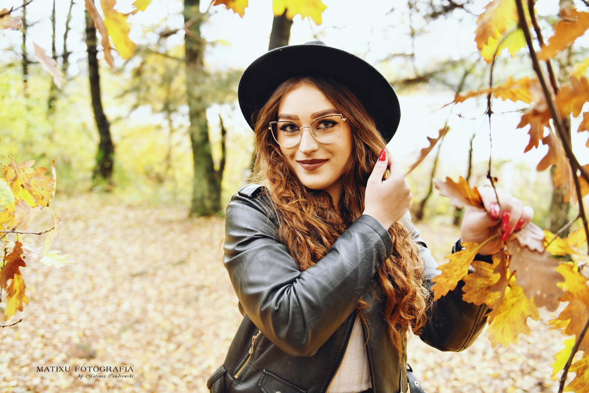 Portret kobiety w czarnym kapeluszu i skórzanej kurtce, pozującej w jesiennym lesie z liśćmi w dłoni