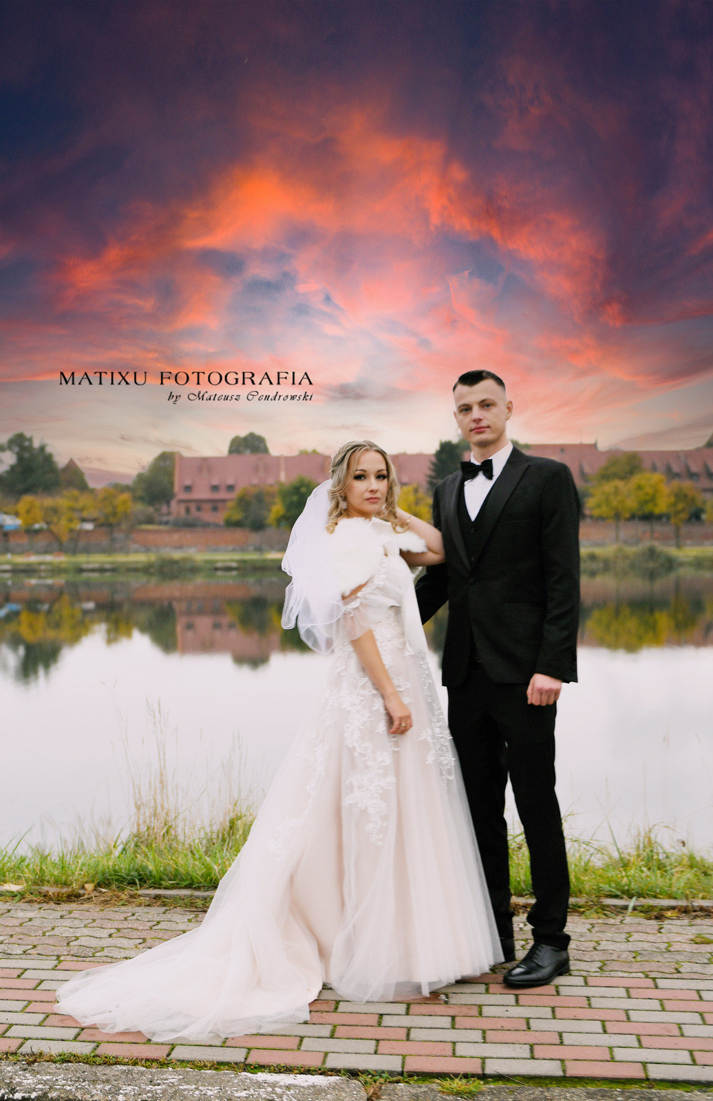 Para młoda: panna młoda w sukni z koronkowymi aplikacjami i futrzanym etolu, pan młody w czarnym smokingu z muszką, pozują na tle jeziora i budynku z czerwonej cegły o zachodzie słońca.
