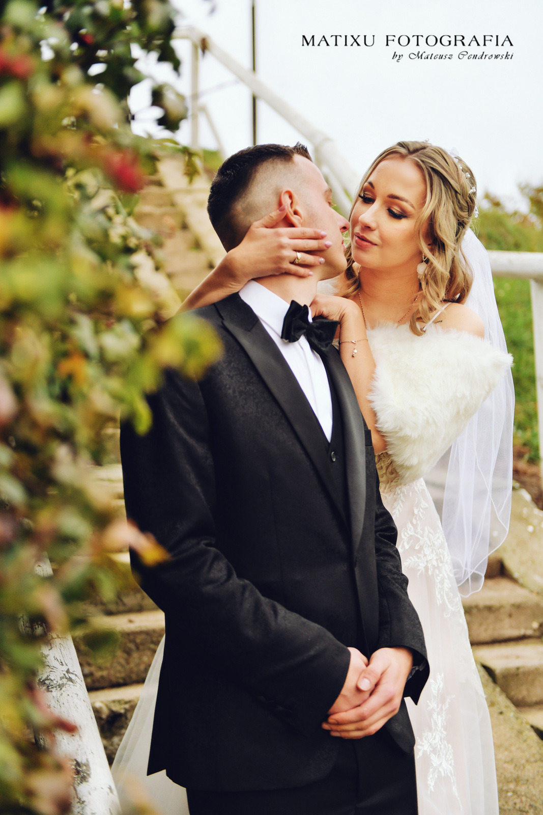 Panna młoda w białej sukni i futrzanym etole dotyka policzka pana młodego w czarnym smokingu na tle schodów i zieleni.