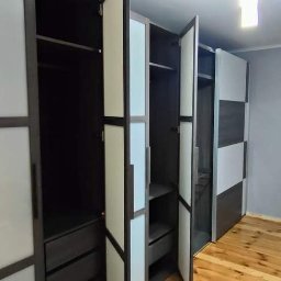 WroPro Meble - Nowoczesna szafa wnękowa z otwartymi drzwiami, widoczne półki i szuflady, w tle okno z żaluzjami i zabezpieczone folią meble.