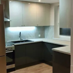 Nowoczesna kuchnia z szarymi szafkami dolnymi i beżowymi górnymi, jasnym blatem, czarnym zlewem i płytkami imitującymi marmur na ścianie, oświetlona punktowymi lampami sufitowymi i listwą LED...