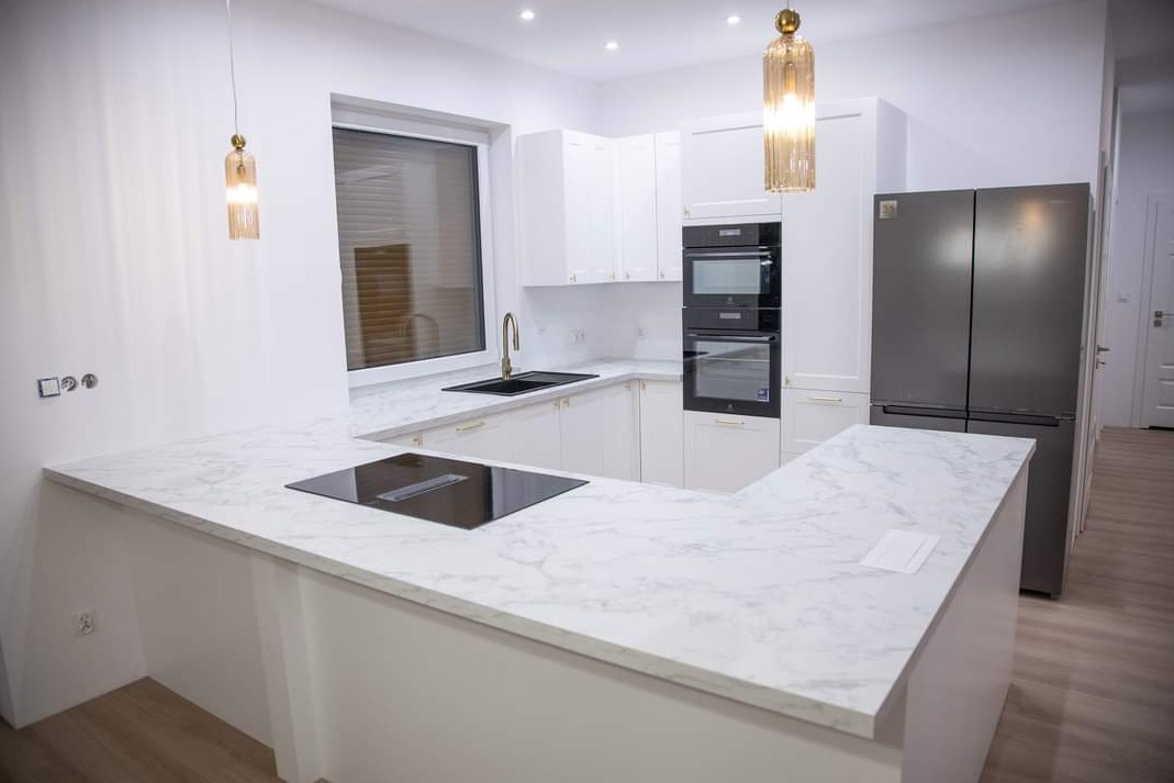 Nowoczesna, jasna kuchnia z białymi szafkami, marmurowym blatem, czarnym zlewem i płytą indukcyjną oraz dużym, dwudrzwiowym, szarym lodówko-zamrażarką.