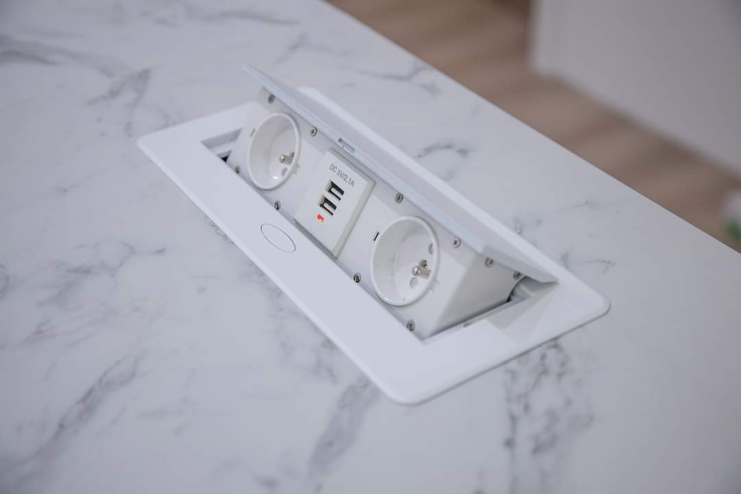 Wpuszczane w blat biurka, wysuwane gniazdo elektryczne z dwoma gniazdami 230V i dwoma portami USB, w białej obudowie, na blacie z imitacją marmuru.