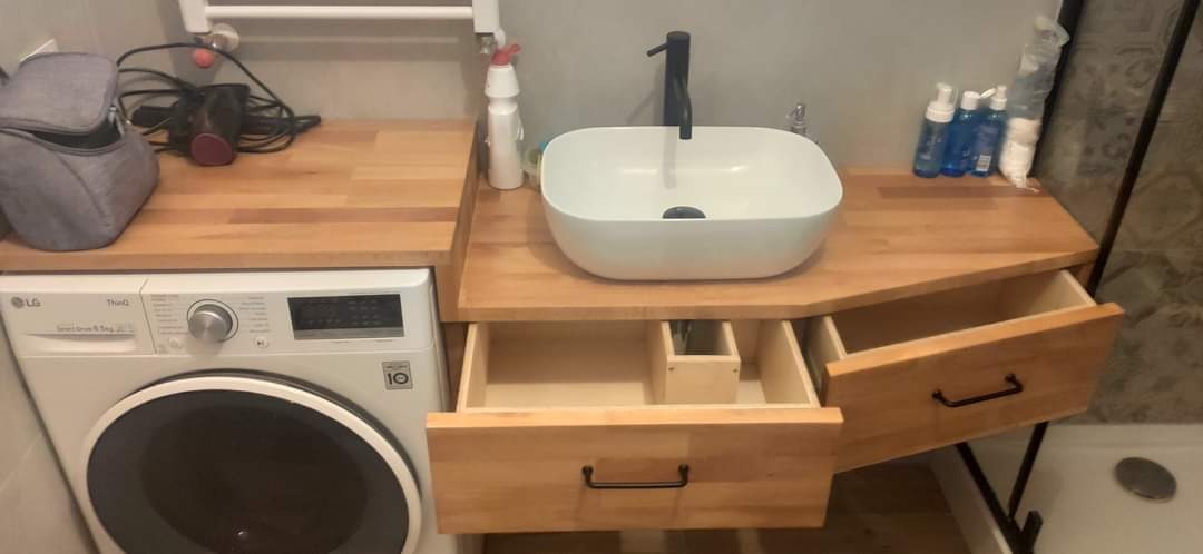 Nowoczesna łazienka z drewnianym blatem, na którym stoi biała umywalka nablatowa z czarną armaturą, obok pralka LG i otwarte szuflady pod umywalką.