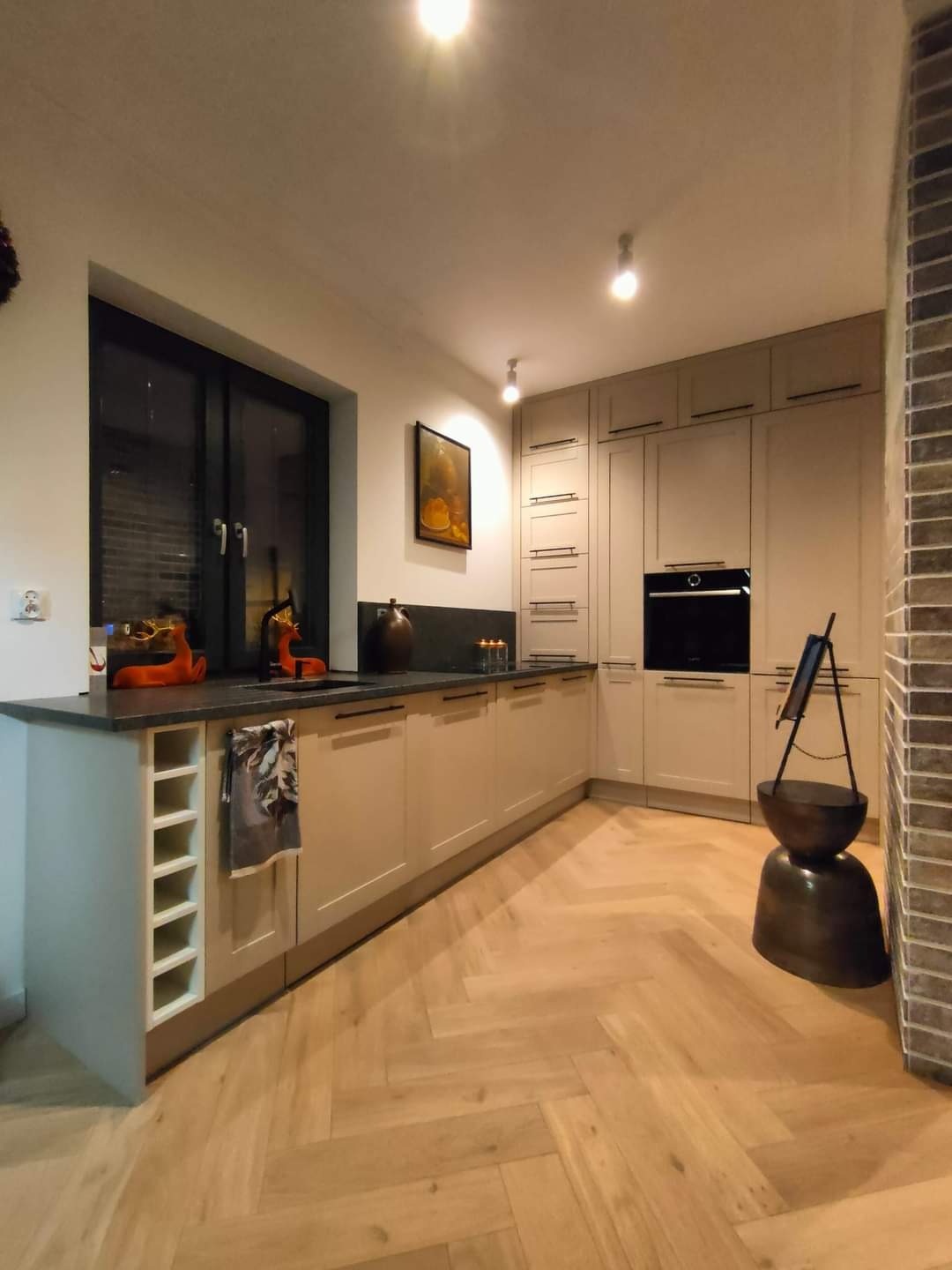 Jasna kuchnia z drewnianą podłogą ułożoną w jodełkę, beżowe szafki z czarnymi uchwytami, czarny blat i okno, dekoracje w oknie i na blacie, nowoczesne oświetlenie.