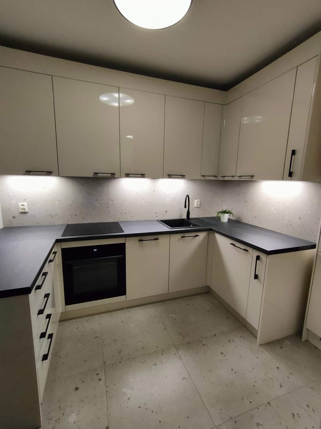 Nowoczesna kuchnia w odcieniach beżu z czarnymi akcentami: blat, armatura, uchwyty. Ściana nad blatem i podłoga w stylu lastryko. Oświetlenie podszafkowe LED. Widok na narożną zabudowę kuchenną ze...