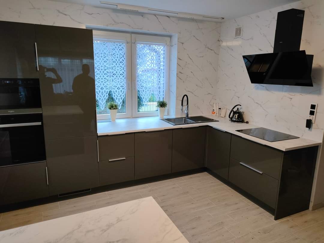 Nowoczesna kuchnia z błyszczącymi, grafitowymi szafkami, białym blatem imitującym marmur, i płytkami ściennymi w tym samym stylu. Widoczny czarny okap i płyta indukcyjna. Na blacie czajnik i drobne...