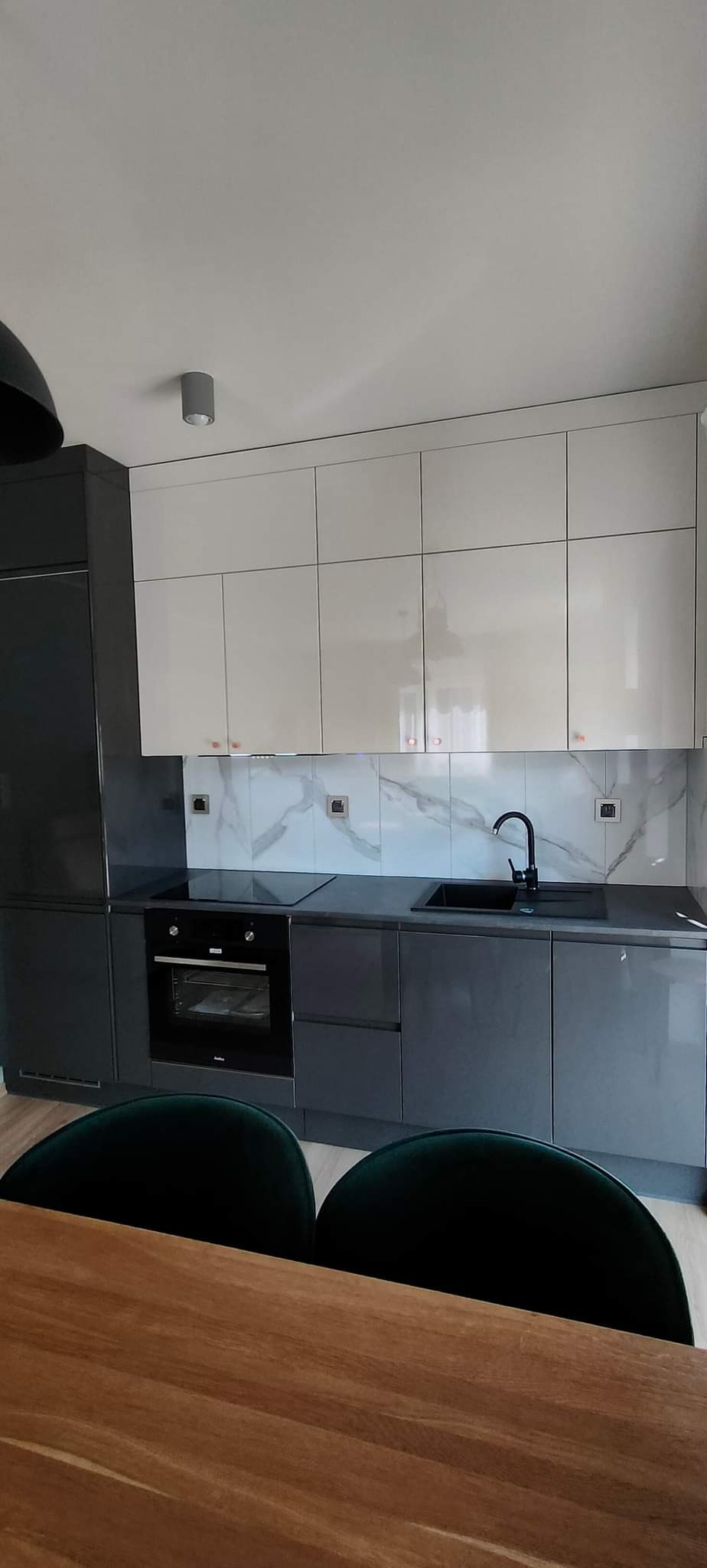 Nowoczesna kuchnia z szarymi szafkami dolnymi i białymi górnymi, marmurowym backsplashiem i czarną armaturą, widok zza drewnianego stołu i zielonych krzeseł.