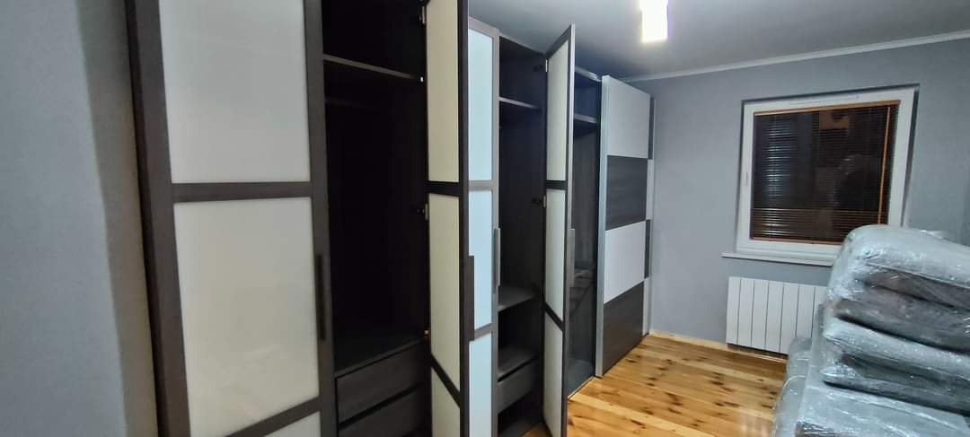 Nowoczesna szafa wnękowa z otwartymi drzwiami, widoczne półki i szuflady, w tle okno z żaluzjami i zabezpieczone folią meble.