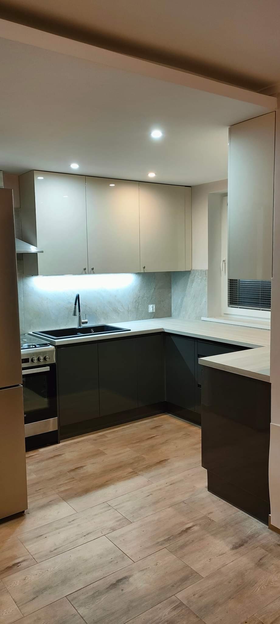 Nowoczesna kuchnia z szarymi szafkami dolnymi i beżowymi górnymi, jasnym blatem, czarnym zlewem i płytkami imitującymi marmur na ścianie, oświetlona punktowymi lampami sufitowymi i listwą LED...