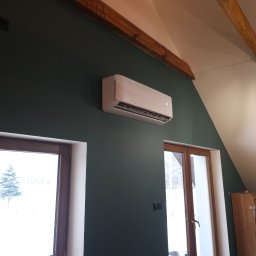 SWPM Szymon Witkowski Performance Management - Biały klimatyzator zamontowany na ciemnozielonej ścianie w pokoju na poddaszu, widoczne drewniane belki stropowe i okna z zimowym krajobrazem.