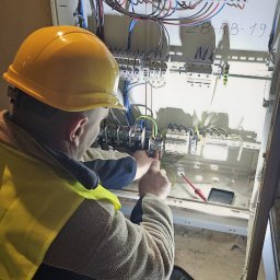 SWPM Szymon Witkowski Performance Management - Elektryk w żółtym kasku i kamizelce, podłącza przewody w skrzynce rozdzielczej, używając śrubokręta; obok rozłożony zestaw narzędzi i dwie akumulatorowe wkrętarki Dewalt.