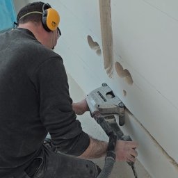 SWPM Szymon Witkowski Performance Management - Elektryk w okularach i słuchawkach ochronnych frezuje bruzdy pod przewody elektryczne w ścianie za pomocą frezarki Metabo z odkurzaczem