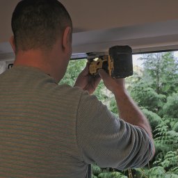 SWPM Szymon Witkowski Performance Management - Elektryk mierzy napięcie w instalacji przy oknie za pomocą miernika zasilanego baterią DeWalt.