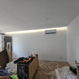 SWPM Szymon Witkowski Performance Management - Wnętrze mieszkania w Łodzi w trakcie montażu klimatyzatora na ścianie, widoczne narzędzia i materiały budowlane, parkiet w jodełkę.