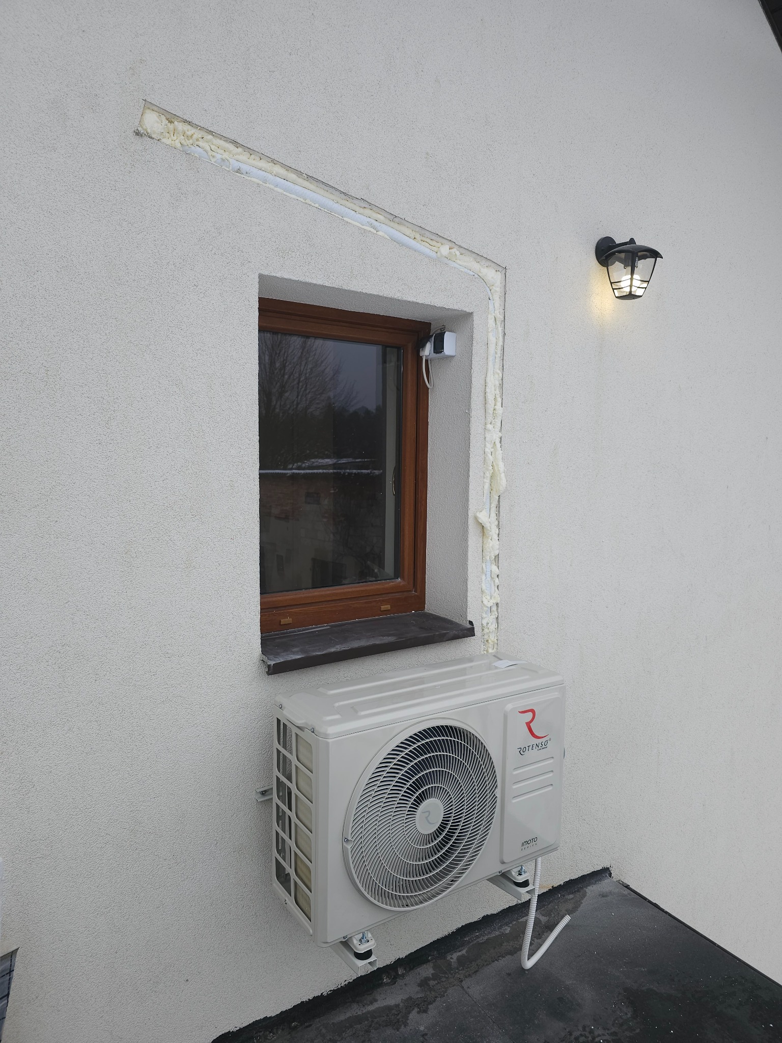 Zewnętrzna jednostka klimatyzacji Rotenso zamontowana pod oknem na elewacji budynku z widocznymi śladami pianki montażowej wokół ramy okiennej.