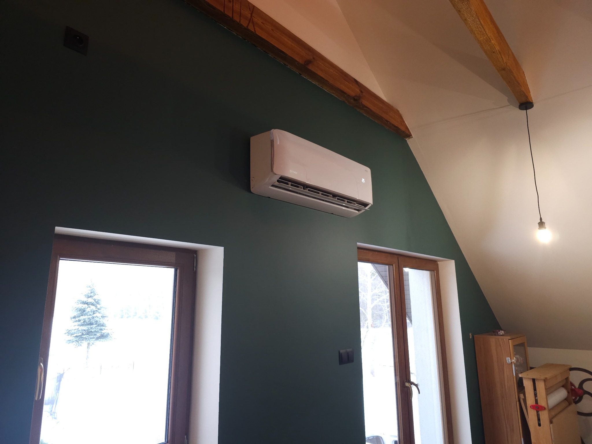 Biały klimatyzator zamontowany na ciemnozielonej ścianie w pokoju na poddaszu, widoczne drewniane belki stropowe i okna z zimowym krajobrazem.
