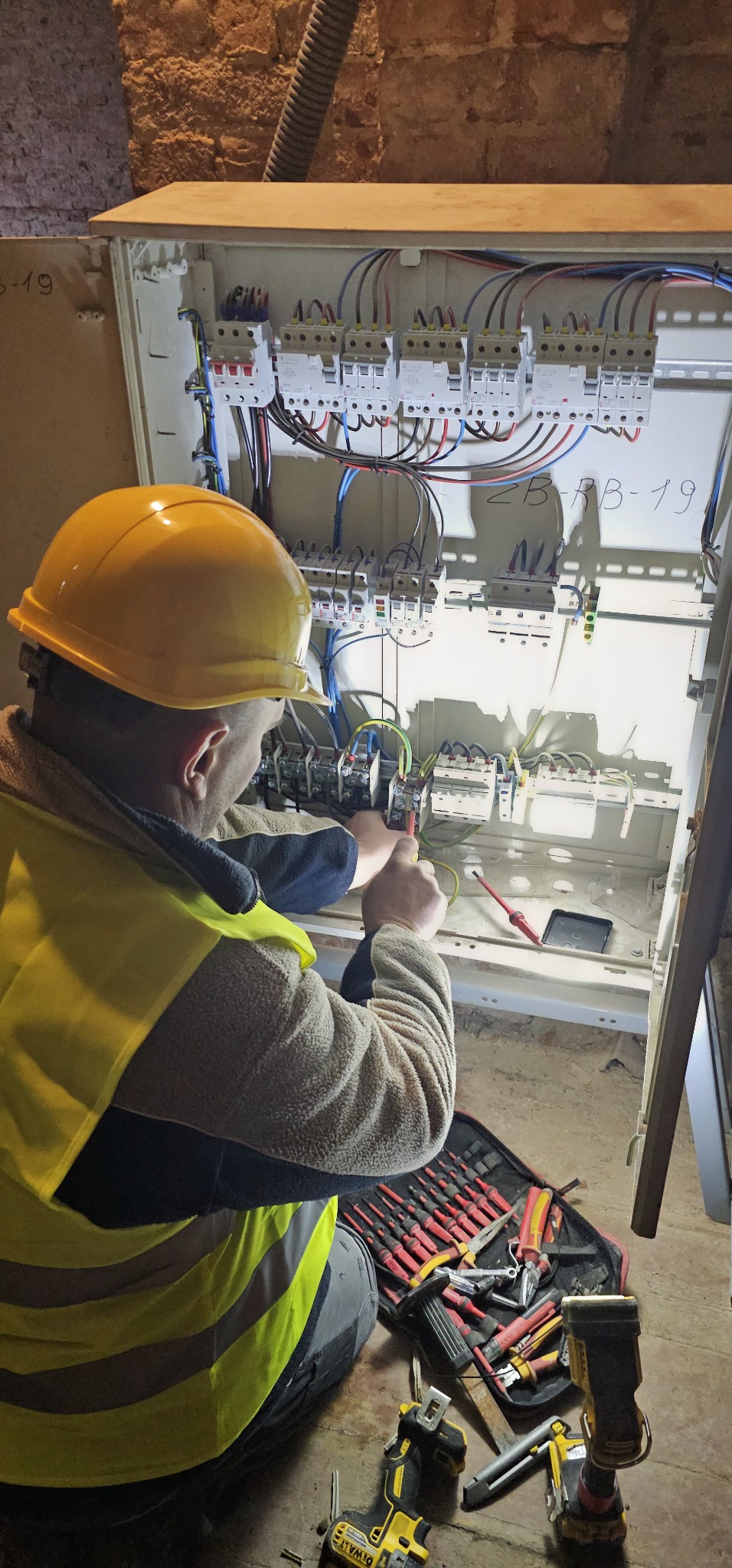 Elektryk w żółtym kasku i kamizelce, podłącza przewody w skrzynce rozdzielczej, używając śrubokręta; obok rozłożony zestaw narzędzi i dwie akumulatorowe wkrętarki Dewalt.