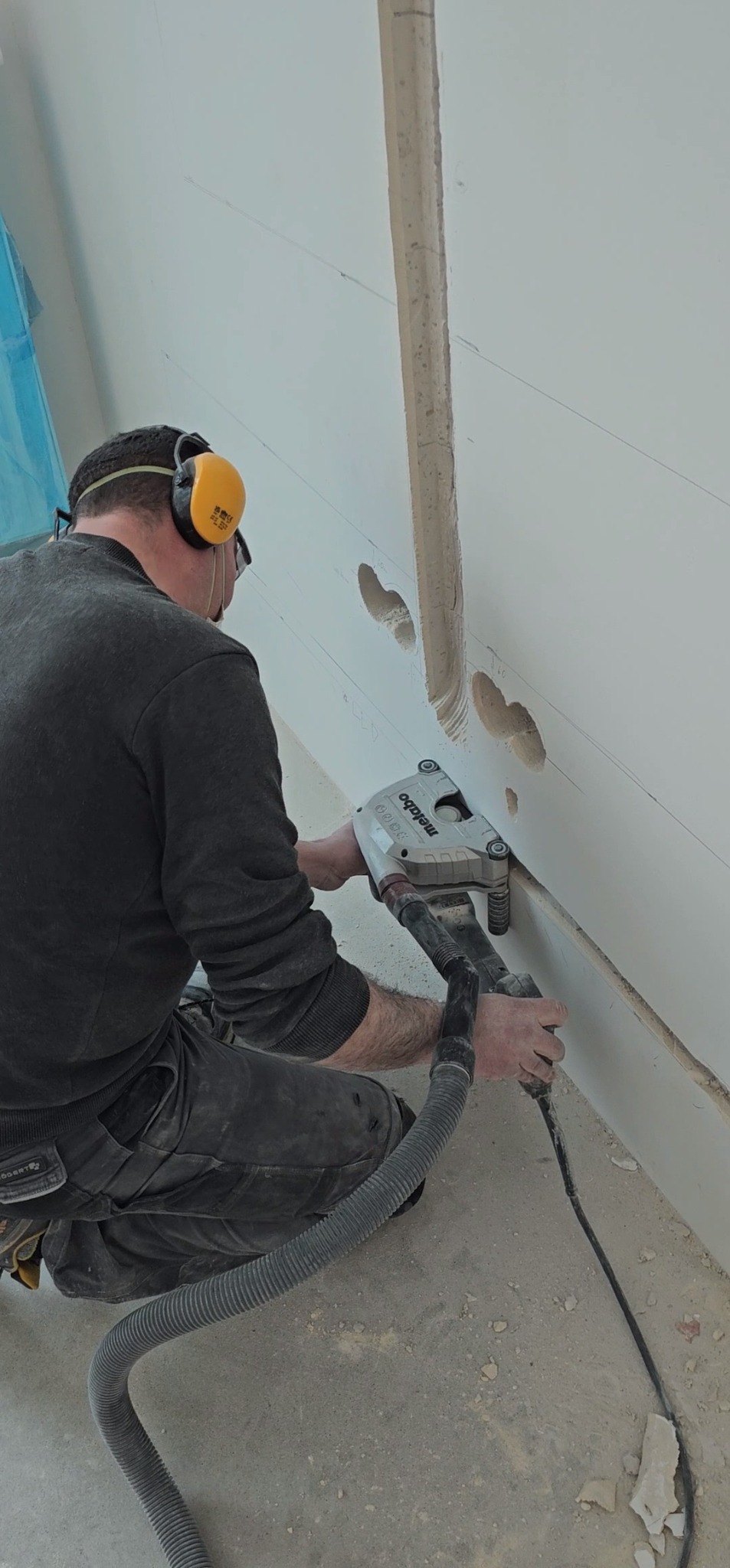 Elektryk w okularach i słuchawkach ochronnych frezuje bruzdy pod przewody elektryczne w ścianie za pomocą frezarki Metabo z odkurzaczem