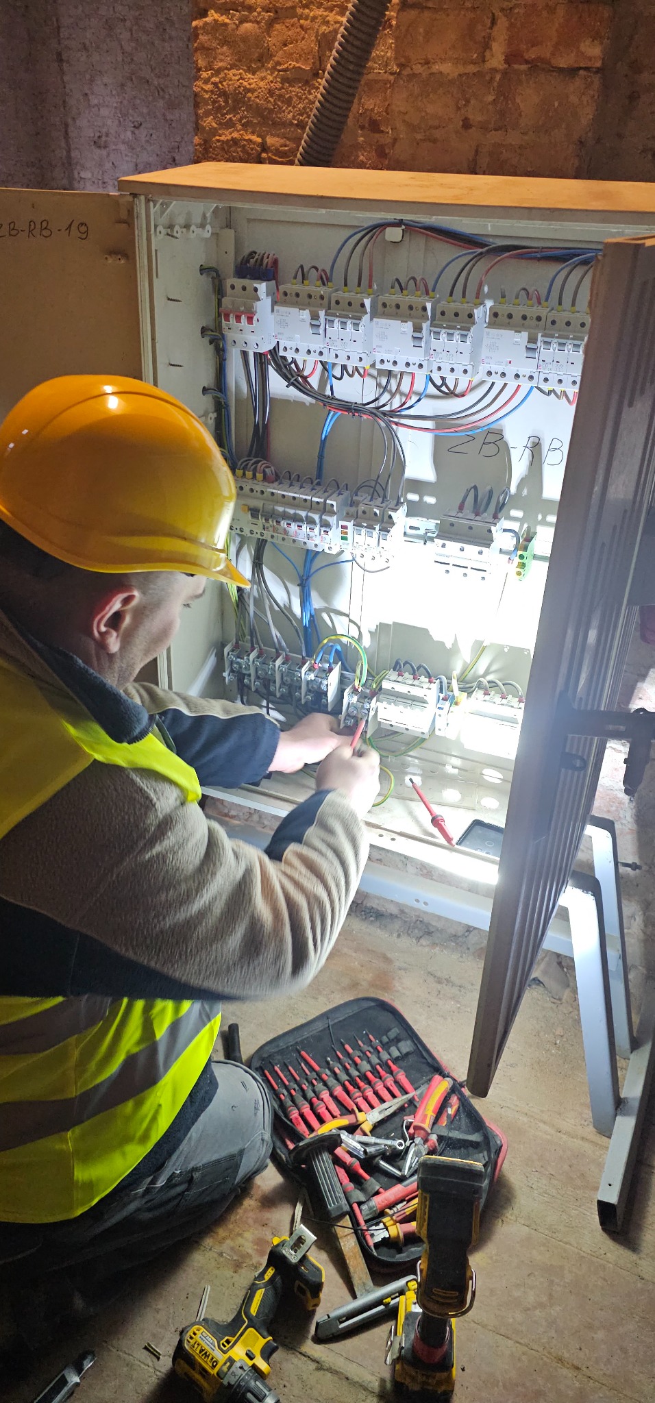 Elektryk w żółtym kasku i kamizelce podłącza przewody w skrzynce rozdzielczej, obok leżą narzędzia: wiertarka, latarka i otwarty futerał z wkrętakami.