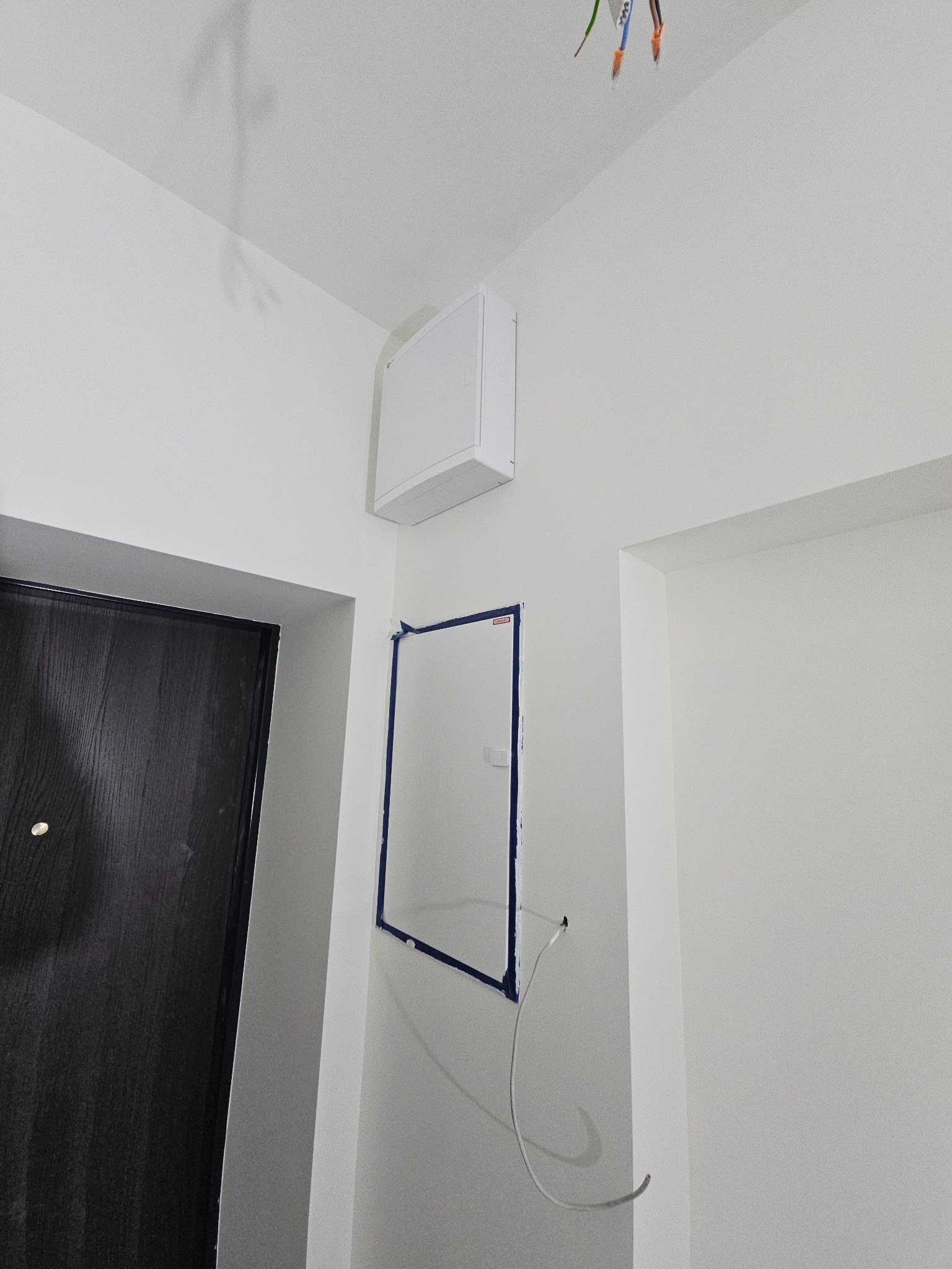 Białe wnętrze z widocznymi przewodami elektrycznymi wystającymi z sufitu, otworem w ścianie zabezpieczonym taśmą malarską i panelem inspekcyjnym na ścianie