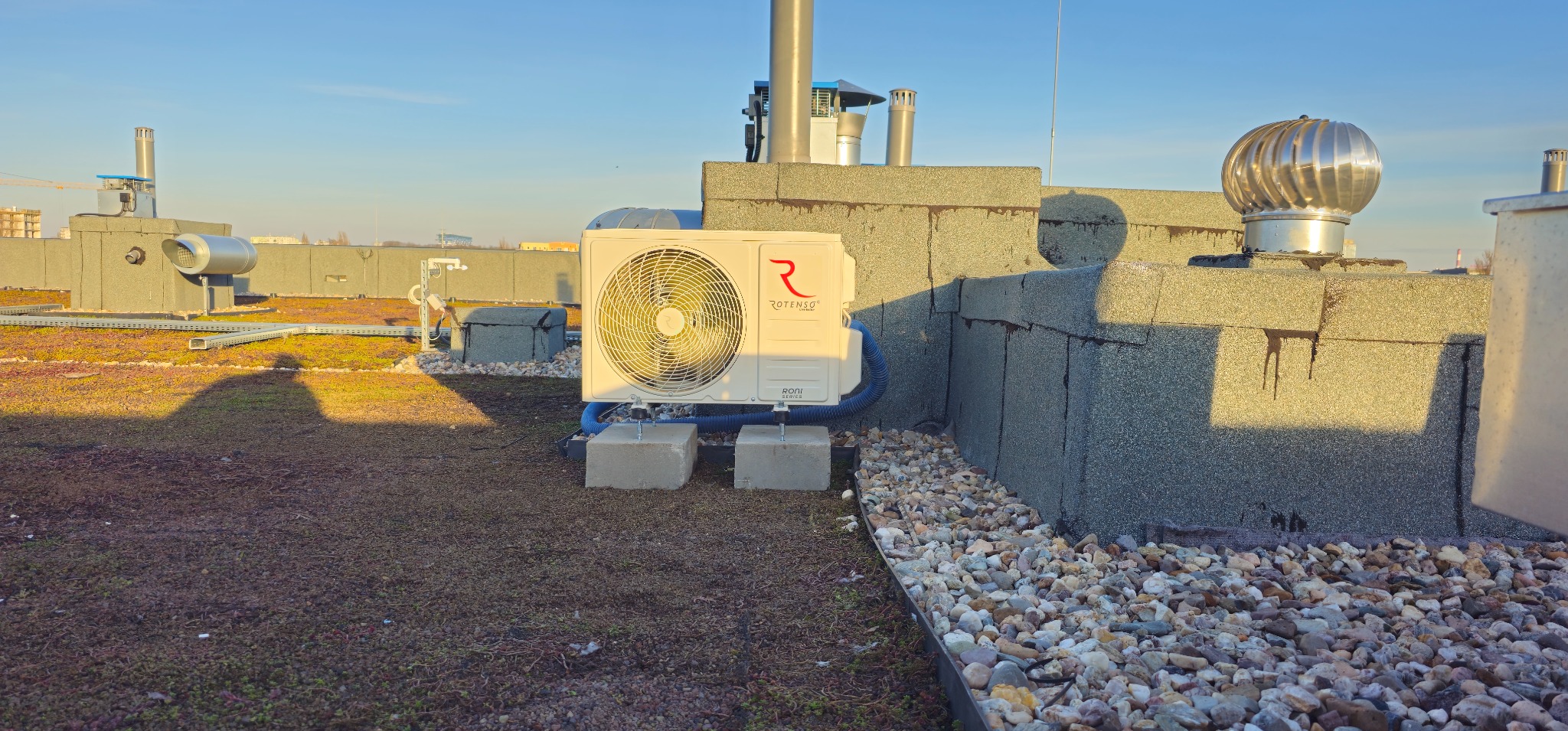 Biały klimatyzator Rotenso Roni Series zamontowany na betonowych blokach na zielonym dachu budynku, z widocznymi elementami wentylacji i kamiennym obrzeżem.