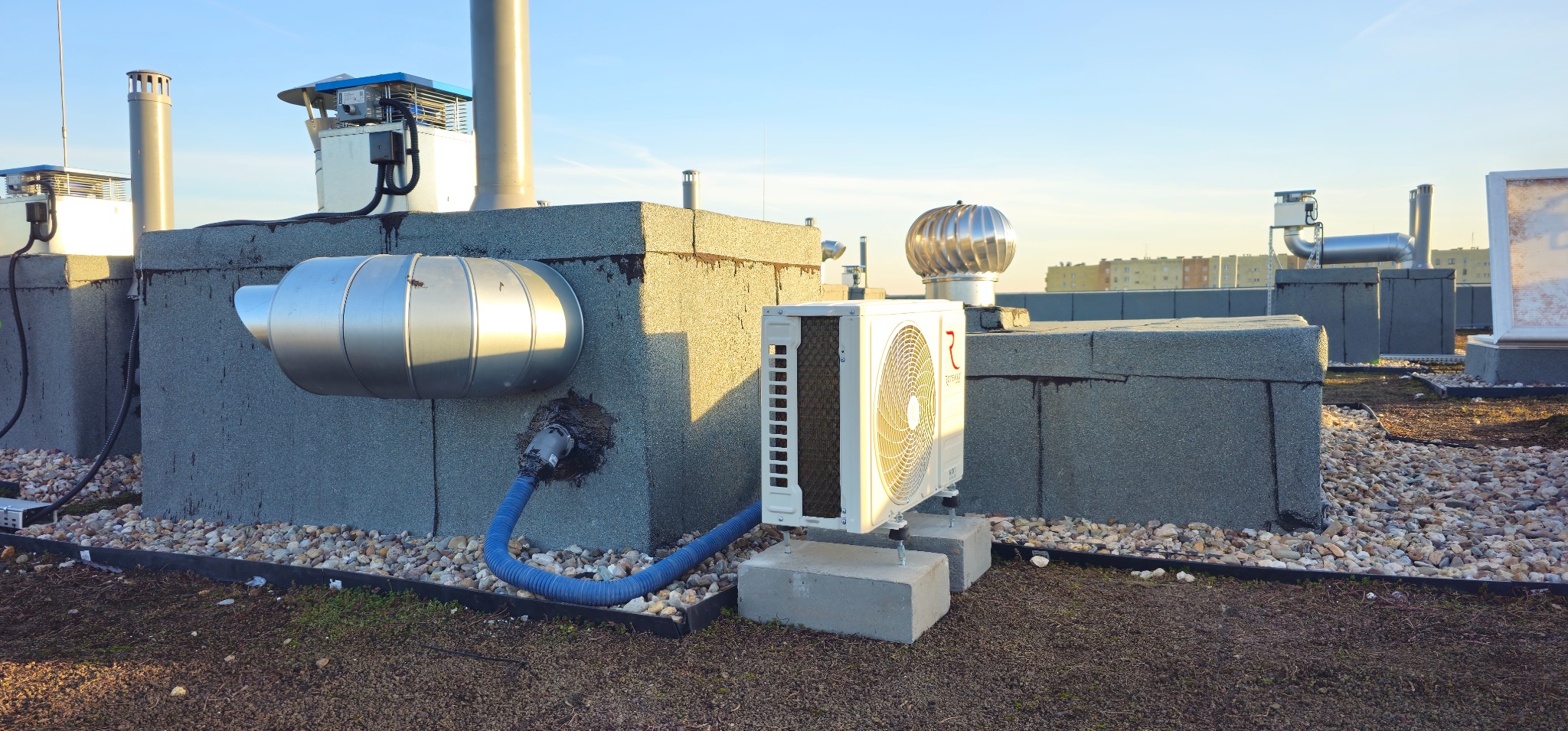 Biały klimatyzator zamontowany na betonowych blokach na dachu budynku, z widocznymi przewodami i innymi instalacjami wentylacyjnymi.