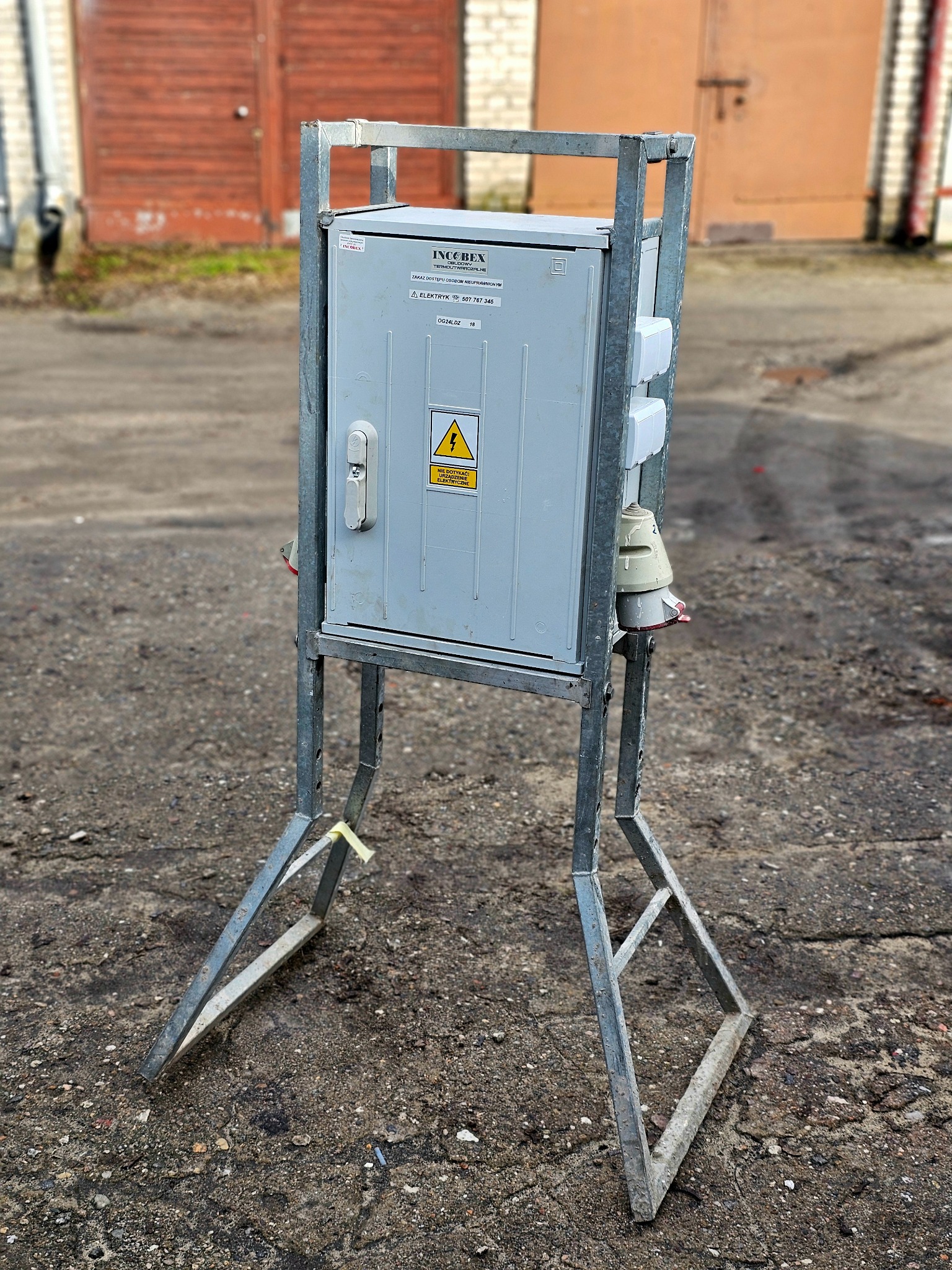 Szara skrzynka elektryczna z ostrzeżeniem o napięciu, zamontowana na metalowym stojaku na zewnątrz, z widocznymi gniazdami i wtyczkami, na tle betonowego placu i dwóch garaży.