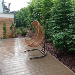 Aura-Wood - Rattanowy fotel wiszący na tarasie z desek kompozytowych, obok zadbany żywopłot i drewniana ścianka z pnączami, widoczne ślady deszczu.