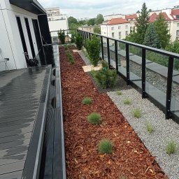 Aura-Wood - Nowoczesny taras na dachu budynku z drewnianą podłogą, rabatami z korą i kamieniami oraz szklaną balustradą, widok na zabudowę miejską.