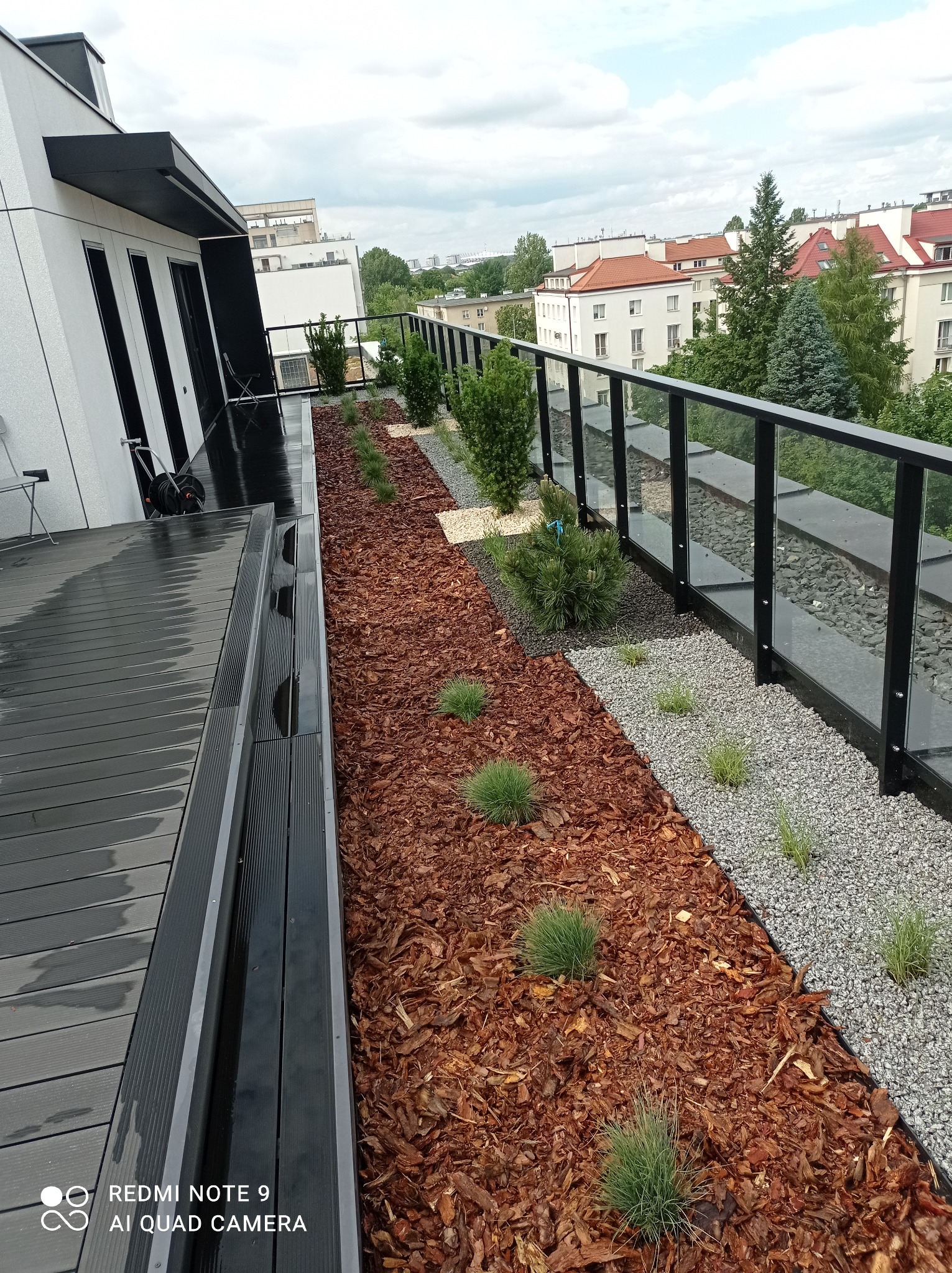 Nowoczesny taras na dachu budynku z drewnianą podłogą, rabatami z korą i kamieniami oraz szklaną balustradą, widok na zabudowę miejską.