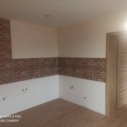 Dawid Gajda - Świeżo wykończone wnętrze z częściowo położoną dekoracyjną czerwoną cegłą na ścianach, widoczne wyprowadzenia elektryczne i hydrauliczne, drewniana podłoga oraz drzwi wewnętrzne z poziomymi frezami.