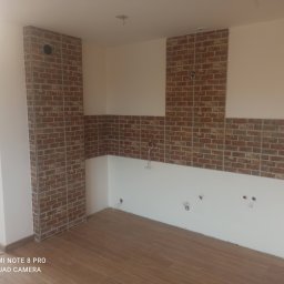 Dawid Gajda - Fragment kuchni w trakcie prac wykończeniowych: ściany częściowo pokryte białą farbą i ceglastymi płytkami, widoczne wyprowadzenia instalacji elektrycznej i wodnej, podłoga wyłożona panelami...