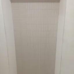 Ściana wykończona jasnymi, pionowo ułożonymi płytkami w kształcie cegiełek, z wąskim pasem płytek poziomych po lewej stronie, otoczona białymi ścianami.